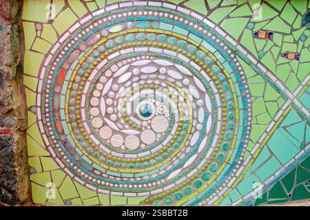 Carreaux de mosaïque colorés représentant des motifs de soleil et de récif Cruz Bay St John USVI | CRUZ BAY, Îles Vierges américaines — Un gros plan d'un panneau mosaïque circulaire avec des motifs de soleil et de récif décore un mur de pierre dans une aire de jeux publique. Des artistes locaux ont construit cette pièce à partir de carreaux de céramique à motifs, de perles de verre et d'incrustations iridescentes disposées en bandes concentriques rayonnant vers l'extérieur. Des mosaïques réalisées par la communauté comme celle-ci sont des éléments courants des espaces publics de la petite ville de Saint-Jean, ajoutant un caractère artistique local à l'infrastructure civique. Banque D'Images