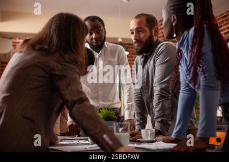 Gros plan de collègues multiethniques debout autour de la table et ayant une conversation importante au bureau. Hommes et femmes d'affaires divers s'appuyant sur le bureau, dans une discussion sérieuse sur le projet de l'entreprise. Banque D'Images