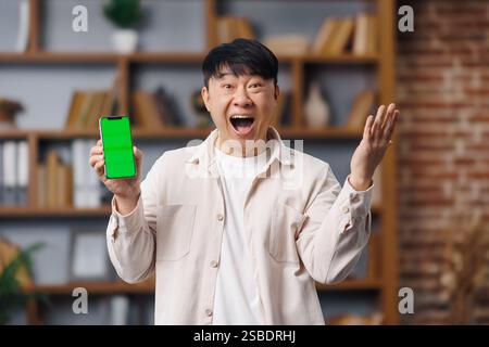 Homme heureux excité, sentiment de gagnant, écran de téléphone, surpris, asiatique, jeune homme présente un smartphone avec un écran vert lumineux dans un intérieur confortable Banque D'Images