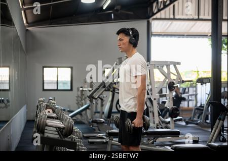 Une vue de côté d'un homme asiatique concentré et déterminé en vêtements de sport et écouteurs travaille avec des haltères devant le porte-haltères et le miroir Banque D'Images