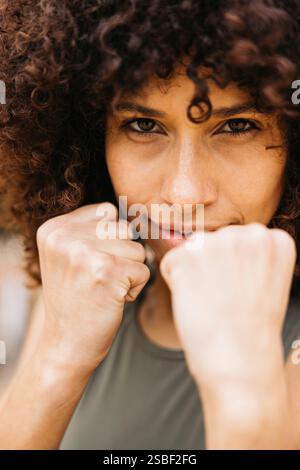 Une jeune femme concentrée avec des cheveux bouclés posant dans une position de combat à l'extérieur, démontrant une forte détermination et une bonne préparation. Son regard intense symboli Banque D'Images