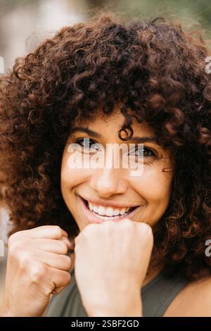 Une femme joyeuse aux cheveux bouclés levant les poings dans une posture de boxe. Elle dépeint la positivité et la détermination. Banque D'Images
