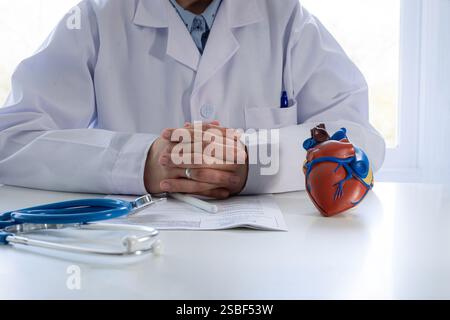 Gros plan d'un médecin méconnaissable dans un manteau blanc, assis à un bureau avec ses mains serrées. Sur la table se trouve un modèle de cœur, un stéthoscope Banque D'Images