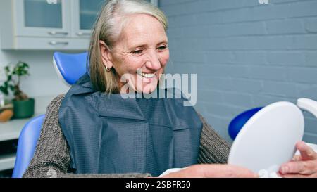 Femme mature souriant à la clinique dentaire tout en vérifiant les dents dans le miroir de main pendant le rendez-vous Banque D'Images