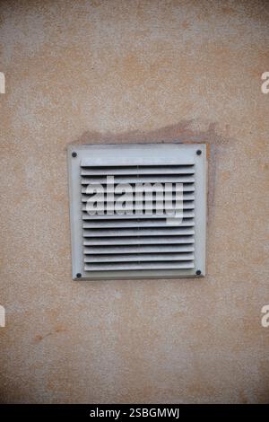 Une grille de ventilation poussiéreuse est montée sur un mur texturé brun clair. Les lattes de la calandre sont légèrement sales, suggérant un certain âge ou un manque de c récent Banque D'Images