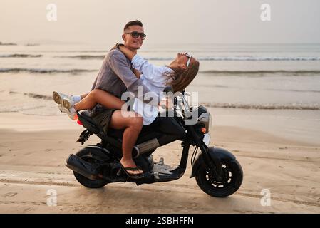 Jeune couple monte en moto par la plage de l'océan. L'homme conduit la moto tandis que la femme se penche joyeusement en arrière. Les amoureux apprécient le voyage romantique en bord de mer en scooter Banque D'Images