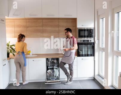 Jeune couple ensemble faisant le ménage dans la cuisine moderne, mettant des assiettes dans le lave-vaisselle Banque D'Images
