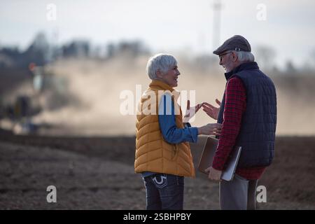 Vue arrière de deux agriculteurs, homme et femme, parlant dans le champ alors que le tracteur labourait en arrière-plan Banque D'Images