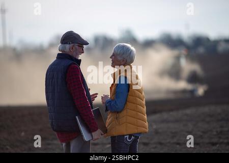 Vue arrière de deux agriculteurs, homme et femme, parlant dans le champ alors que le tracteur labourait en arrière-plan Banque D'Images