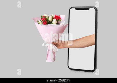 Main avec bouquet de belles fleurs sortant de l'écran du smartphone sur fond gris Banque D'Images