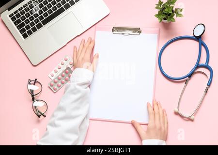 Médecin féminin avec presse-papiers vierge, stéthoscope et ordinateur portable sur fond rose. Journée mondiale de la santé Banque D'Images