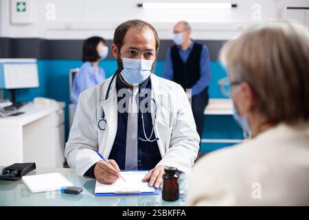Médecin masculin explique le formulaire d'évaluation de la santé à la femme senior pendant le rendez-vous clinique. Le médecin masqué écoute attentivement la patiente retraitée et écrit ses symptômes sur le presse-papiers. Banque D'Images