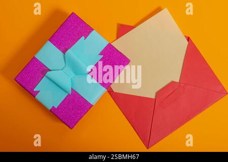 Boîte cadeau brillante colorée lumineuse décorée avec un noeud en ruban près de l'enveloppe ouverte sur fond orange vif. Parfait pour les célébrations, les cadeaux ou les événements Banque D'Images