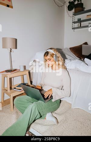 Jeune étudiante avec des écouteurs sans fil à l'aide d'un ordinateur portable assis près du lit dans la chambre de dortoir de l'université Banque D'Images