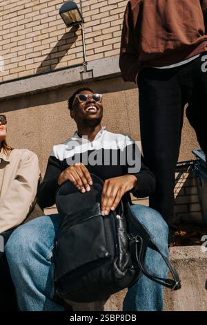 Vue à angle bas de souriant étudiant masculin ouvrant sac à dos tout en étant assis avec des amis sur le campus de l'université Banque D'Images