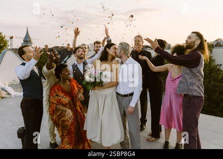 Heureux invités de mariage jetant des confettis sur le couple nouvellement marié embrassant sur le toit pendant la célébration de mariage Banque D'Images
