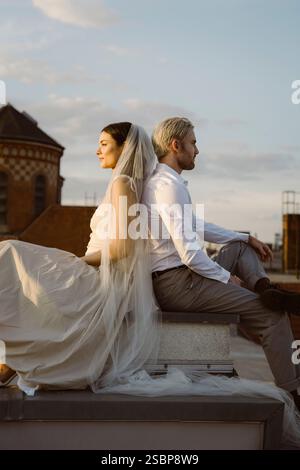 Vue latérale d'un jeune couple multiracial nouvellement marié assis dos à dos sur le toit contre le ciel Banque D'Images