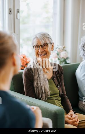 Femme âgée souriante assise sur le canapé et parlant avec le soignant à la maison de retraite Banque D'Images