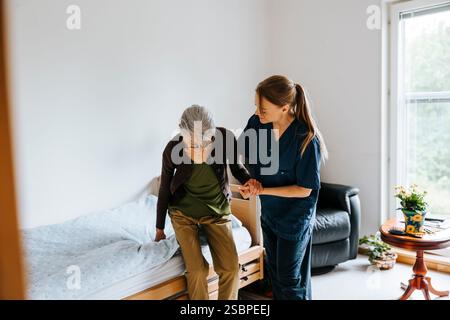 Femme gardienne aidant une femme âgée assise sur le lit à la maison de retraite Banque D'Images