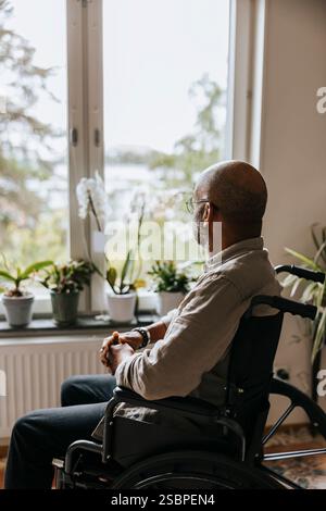 Homme âgé solitaire regardant par la fenêtre alors qu'il était assis en fauteuil roulant à la maison de retraite Banque D'Images