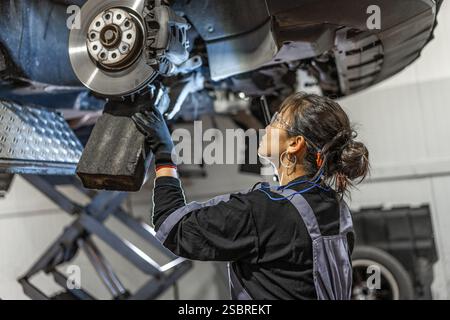 Mécanicien féminin qualifié travaillant sous une voiture levée, inspectant et réparant le système de freinage dans un atelier professionnel Banque D'Images