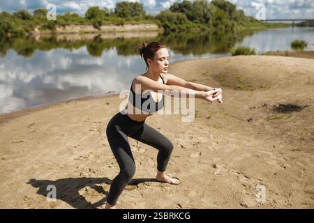 Femme effectue un exercice de squat sur une rive sablonneuse, tenant une bande de résistance Banque D'Images