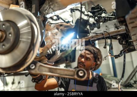 Jeune mécanicien utilisant la lampe de poche inspectant le châssis et la suspension de voiture dans un atelier de réparation automobile professionnel Banque D'Images