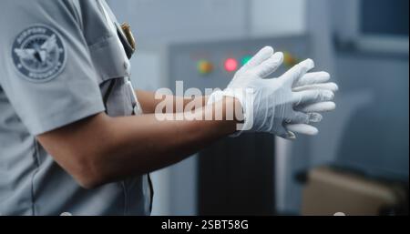 Point de contrôle de sécurité de l'aéroport : valise se déplaçant dans le scanner de détecteur de métaux à rayons X avancé. Focus sur l'agent de sécurité afro-américain portant des gants se préparant aux procédures de contrôle des bagages de la TSA. Banque D'Images