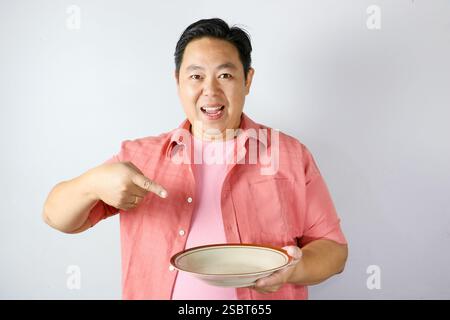 Un homme portant une chemise rose avec un visage joyeux souriant et pointant son doigt sur une assiette à manger vide marron. Banque D'Images