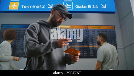 Terminal de l'aéroport occupé : l'homme avec sac à dos vérifie les informations de vol en scannant le billet à l'aide du téléphone. Voyageur partant en voyage de vacances. Panneau numérique d'arrivée et de départ pour les passagers en arrière-plan. Banque D'Images