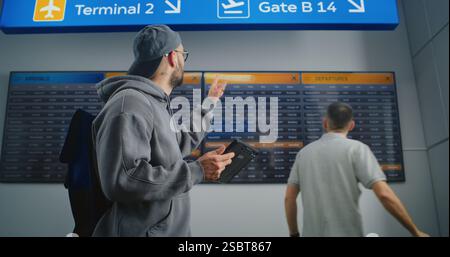 Terminal de l'aéroport occupé : le voyageur avec sac à dos utilise une tablette, vérifie les informations de vol sur le panneau de départ numérique, part en voyage de vacances. Affichage des horaires de l'aéroport pour les passagers en arrière-plan. Banque D'Images