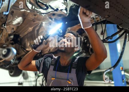 Jeune mécanicien utilisant une lampe de poche, inspectant le dessous du véhicule soulevé sur un ascenseur dans un atelier de voiture Banque D'Images