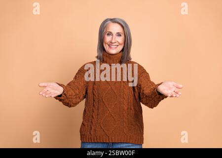 Charmante femme senior souriant chaleureusement, portant un élégant pull marron tout en posant à bras ouverts dans un geste de bienvenue Banque D'Images