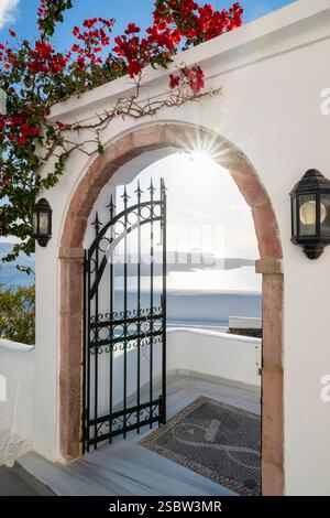Vue sur la caldeira de Santorin et le soleil à travers une arche avec une porte ouverte en fer forgé et bougainvilliers en fleurs, Cyclades, Grèce Banque D'Images