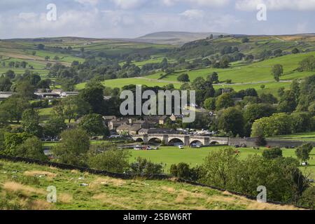 Burnsall niché dans la vallée (pont voûté, maisons et chalets, champs verdoyants à flanc de colline, vue longue distance, été) - Yorkshire Dales, Angleterre, Royaume-Uni. Banque D'Images