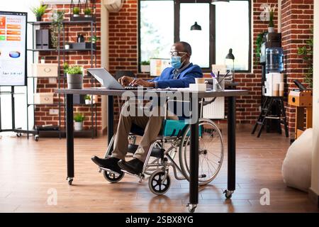 Afro-américain utilisateur masculin en fauteuil roulant portant un masque facial et tapant sur ordinateur portable, mettant à jour les informations de l'entreprise. Homme d'affaires recherchant des stratégies de marketing sur l'appareil au bureau dans le bureau de démarrage. Banque D'Images