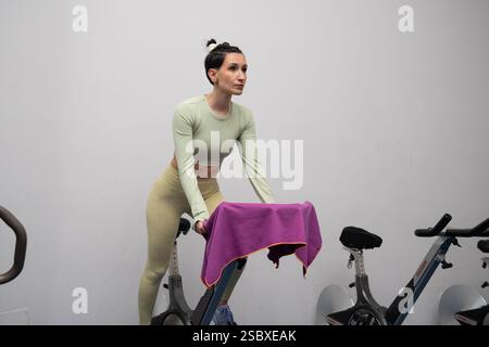 Jeune femme vêtue de vêtements de sport élégants, ajustant le vélo de rotation avec une serviette violette vibrante, se préparant pour une séance d'entraînement énergisante à Banque D'Images