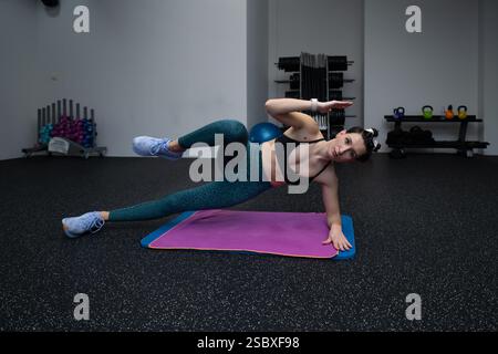 Jeune sportive pratiquant la variation de pose de planche latérale sur tapis violet dans le centre de fitness, renforçant les muscles du tronc et favorisant la stabilité avec focuse Banque D'Images