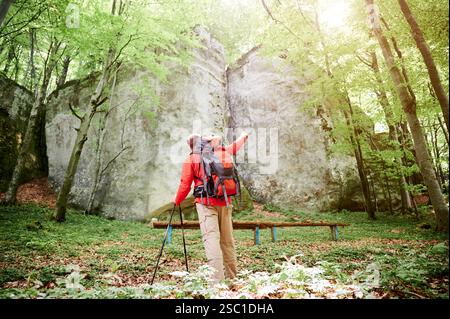 La vue arrière du randonneur avec un grand sac à dos pointe vers une formation rocheuse imposante dans une forêt luxuriante et ensoleillée. Avec des bâtons de trekking à la main, les touristes se dressent au milieu d'un feuillage vert vibrant et d'un soleil tamisé. Banque D'Images