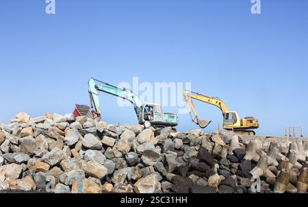 Deux excavatrices travaillent avec diligence sur un chantier de construction tétrapode au niveau de brise-lames entourés de gros rochers sous un ciel bleu clair et lumineux Banque D'Images