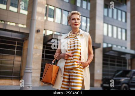 Femme d'affaires mature élégante avec les cheveux courts dans un gilet blanc élégant Workwear, pantalon rayé jaune et blanc et collier de perles, debout à l'extérieur en C. Banque D'Images