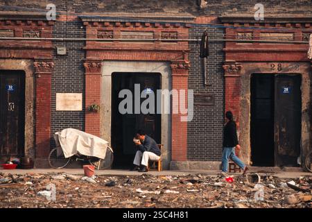 Des gens devant des bâtiments en briques dans une rue en Chine, avec quelques débris visibles sur le sol. Banque D'Images