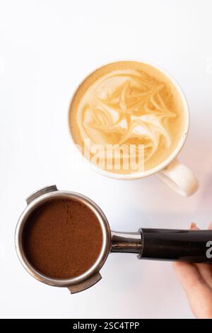 Vue de dessus d'un porte-filtre avec café moulu et d'une tasse de latte avec permis Main du barista tenant l'équipement sur fond blanc. Banque D'Images