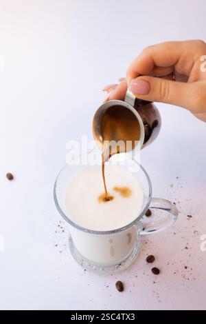Main féminine versant un espresso frais à partir d'une dose de métal dans un verre de lait, faisant un latte ou un cappuccino. Processus de fabrication de café sur fond blanc. Banque D'Images