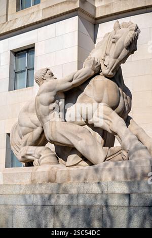 Homme contrôlant le commerce, sculpture au bâtiment de la Federal Trade Commission à Washington DC | WASHINGTON — Un groupe sculpté en calcaire intitulé « Homme contrôlant le commerce » saisit un cheval cabré au coin du bâtiment de la Federal Trade Commission à Washington, D.C. Sculpté par Michael Lantz, l’œuvre était l’une des deux groupes flanquants créés pour l’apex oriental du bâtiment, une allégorie du gouvernement réfrénant le pouvoir du monopole. Lantz remporta le concours « Apex » de la section du Trésor de 1937-38 à l’âge de 29 ans, inconnu, puis sculpta la paire sur place avant sa révélation en 1942. Banque D'Images