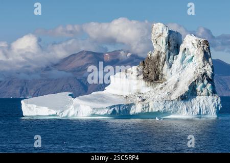 Un iceberg couvert de débris du glacier flotte dans la baie de Baffin près de la côte ouest du Groenland avec ses montagnes couvertes de glacier. Groenland, Danemark Banque D'Images