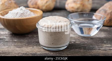 Une entrée pétillante au levain dans un bocal en verre, entourée de farine, d'eau et de pain fraîchement cuit sur une table en bois rustique. Banque D'Images