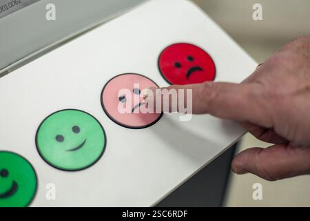 Main appuyant sur un bouton emoji légèrement insatisfait sur une machine de rétroaction de satisfaction de la clientèle avec des smileys verts, roses et rouges, exprimant un neutre Banque D'Images