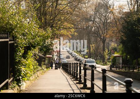 Piétons et voitures par une journée ensoleillée dans la rue de Primrose Hill, Londres, Royaume-Uni. La circulation coule le long de la route bordée d'arbres. Banque D'Images