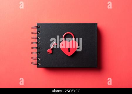 Cadenas rouge en forme de coeur avec une clé sur un cahier noir sur un fond rouge. Vue de dessus, pose à plat. Banque D'Images